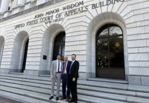 Three men in business suits pose on the stone steps outside a courthouse building with arched windows behind them.