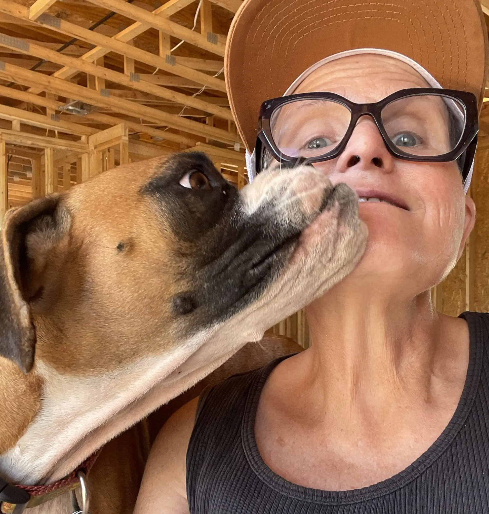 Close-up of a person wearing a brown sun hat and large black glasses while a dog licks their cheek; wooden framing visible in the background.