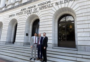 Three men in business suits pose on the stone steps outside a courthouse building with arched windows behind them.