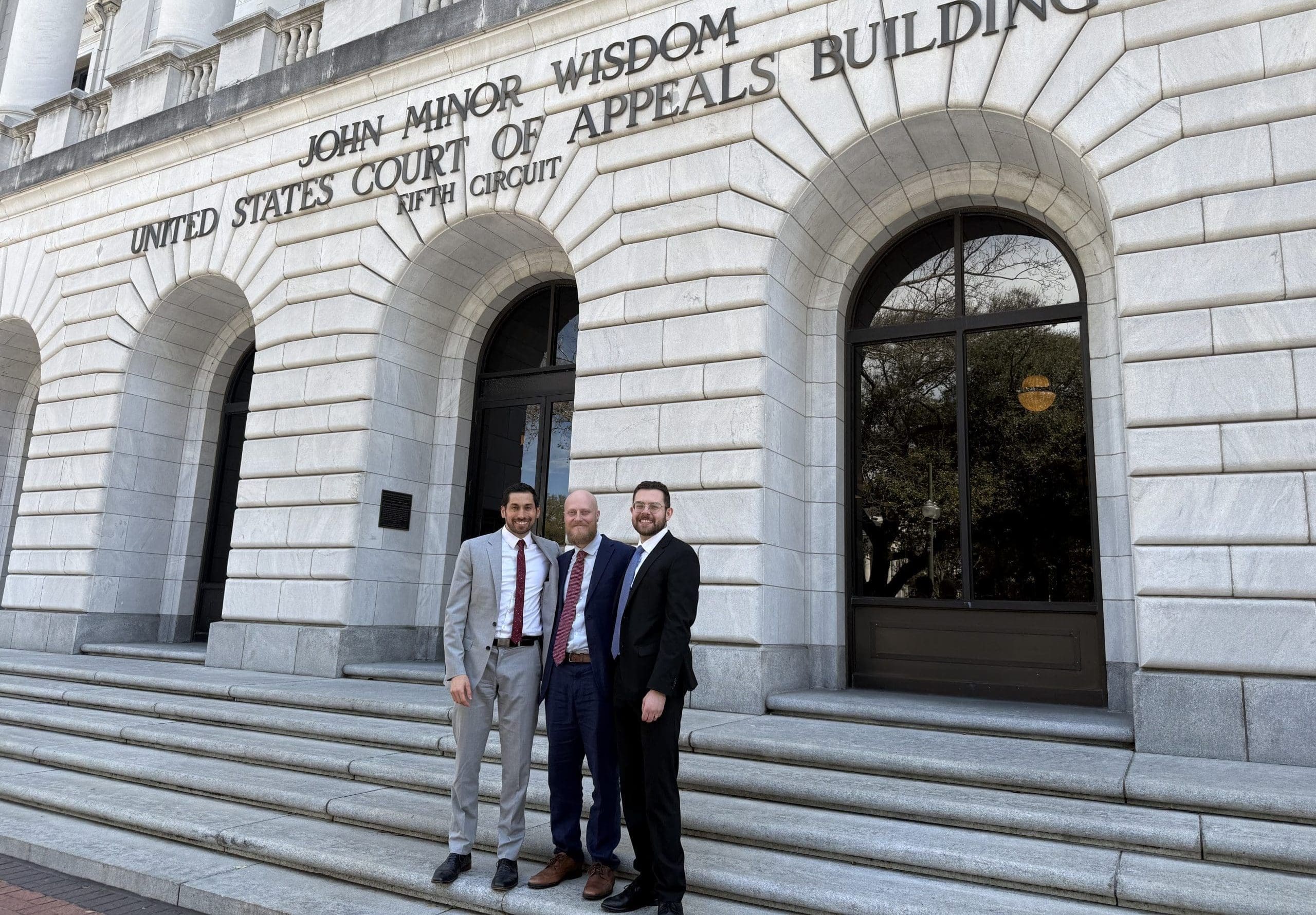 Three men in business suits pose on the stone steps outside a courthouse building with arched windows behind them.