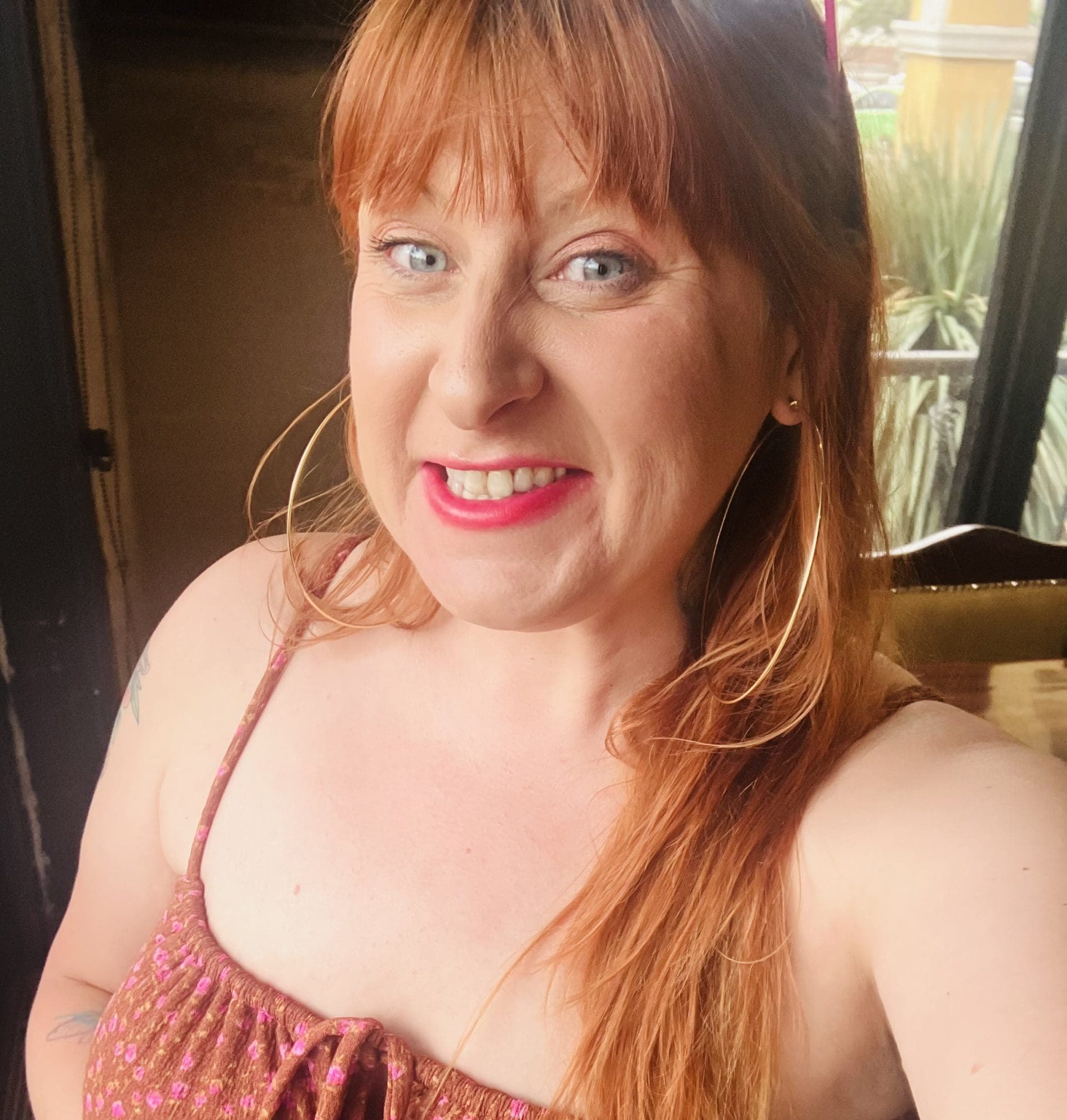 Close-up of a smiling woman with long red hair wearing a brown patterned dress, standing by a doorway.